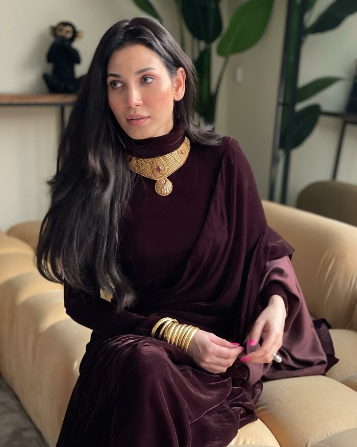 Woman in maroon outfit and gold jewelry sitting on a beige sofa, relating to intoxicated man's threat and supermodel wife defense. Woman in maroon outfit and gold jewelry sitting on a beige sofa, relating to intoxicated man's threat and supermodel wife defense.