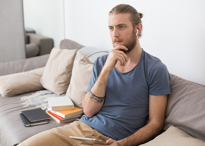 A young man sitting on a couch, wearing earbuds, looking thoughtful while holding a tablet, reflecting on rich people moments. - 16