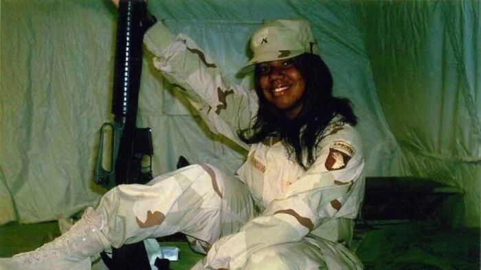 Smiling soldier in desert camouflage uniform holding rifle inside military tent, unsettling image from Morbid Knowledge group.