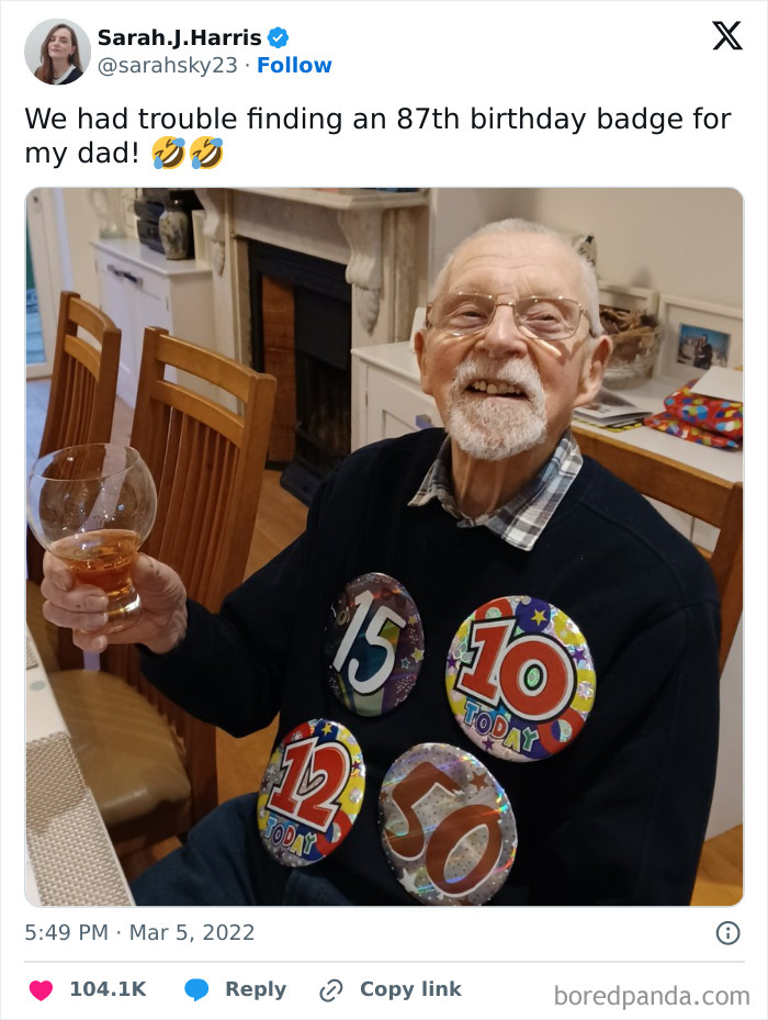 Elderly man wearing multiple birthday badges, smiling and holding a glass in a home setting, British memes humor.