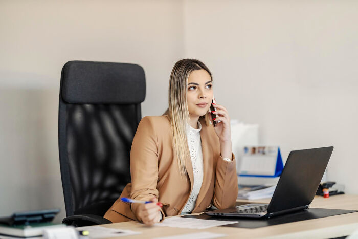 Young woman in a tan blazer using phone and laptop in office applying unhinged hacks to land job successfully.