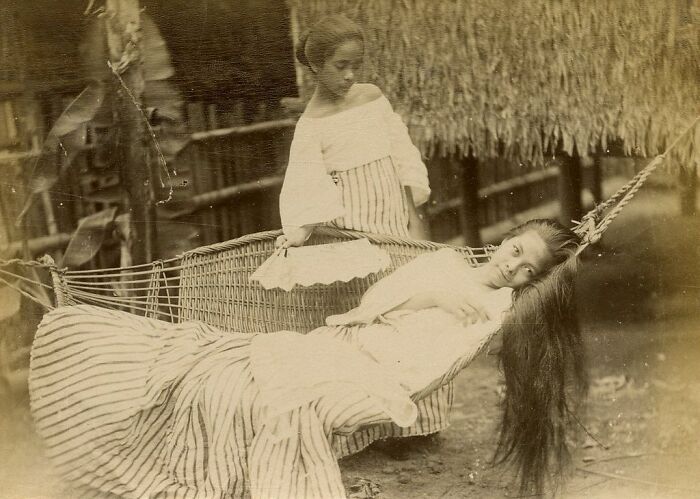 Two Filipino women in traditional clothing resting on a hammock under a nipa hut in the Philippines in the 1890s.