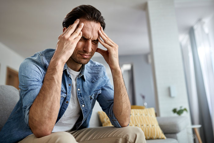 Man sitting on couch with hands on temples, stressed and frustrated in a modern living room setting.