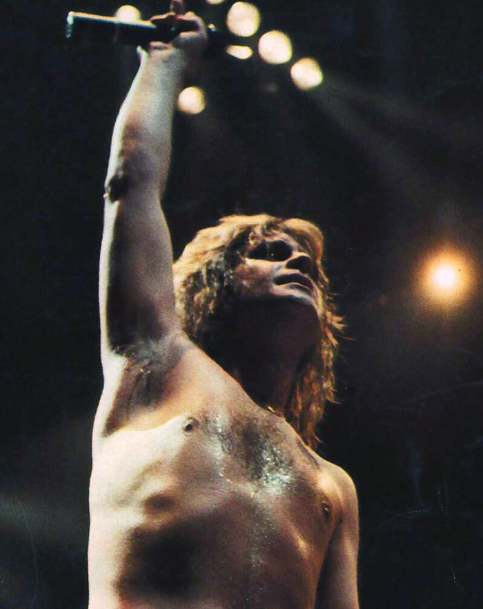 Shirtless male rock performer on stage holding microphone up, showcasing insane family flexes with intense lighting.