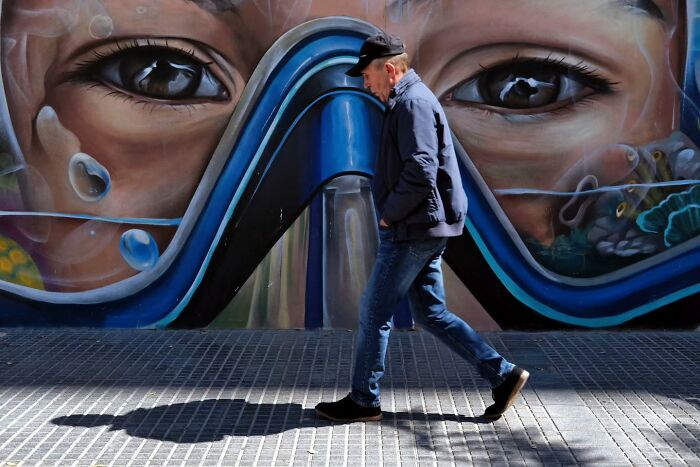 Man walking on street in front of large mural, capturing unseen unposed moments of life on the streets.