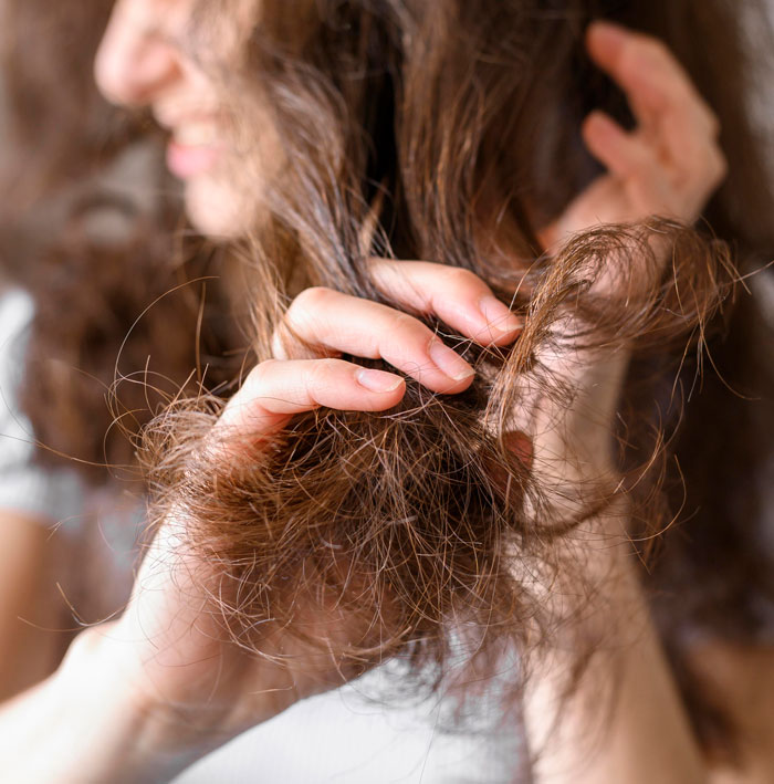 Woman blaming sexism for job rejection, holding and examining her damaged hair with blurred face in the background.