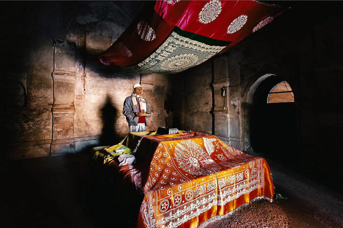 Man in traditional clothing standing in a dimly lit room with colorful fabric, capturing candid everyday moments from Asia.