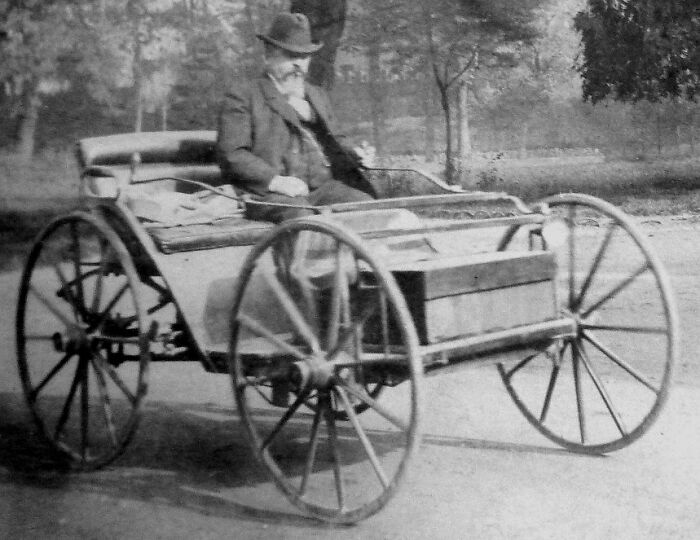 Early 20th century photo of a man driving a wild first car with large wooden wheels on a dirt road.