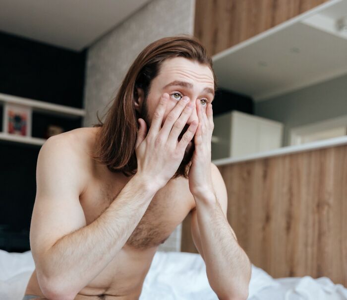 Shirtless man with long hair sitting on bed, covering face with hands, showing feelings after NSFW experience shared online.