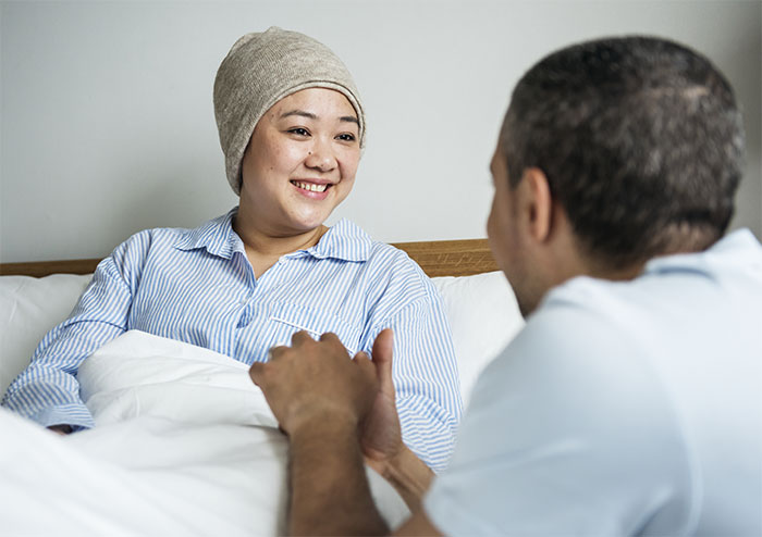 Woman in hospital bed wearing headscarf, smiling and holding hands with a man, reflecting on marriage and care challenges.