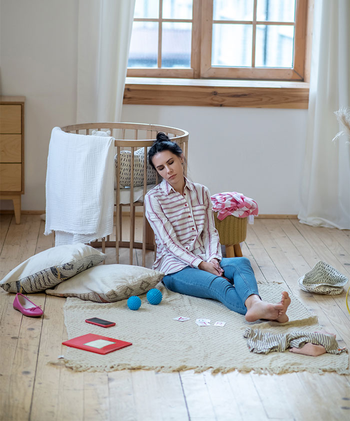 Tired woman sitting on floor near baby crib, upset about cousin refusing to babysit instead of attending school.