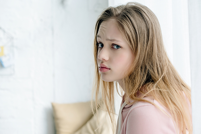 Teenage woman with a sad expression sitting by a window, reflecting on family and affair baby issues.