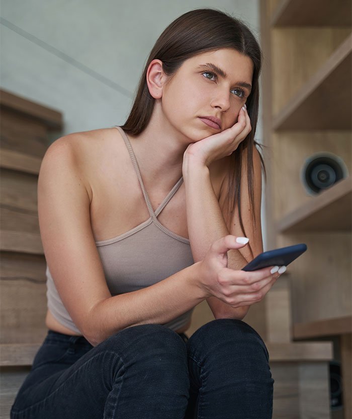 Young woman sitting alone at home looking pensive, holding phone, representing babysitter and mom's Halloween party dilemma. - 8