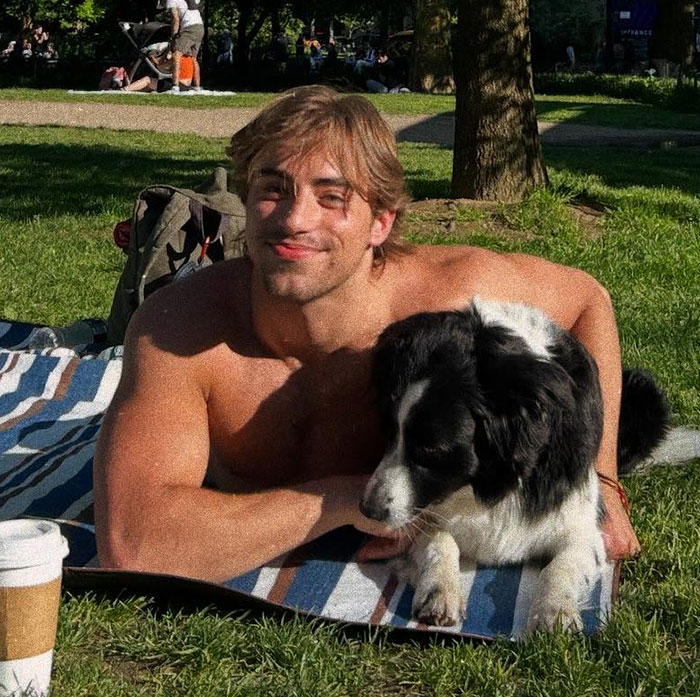 Young man lying shirtless on a blanket outdoors, smiling with a black and white dog by his side in a park setting.