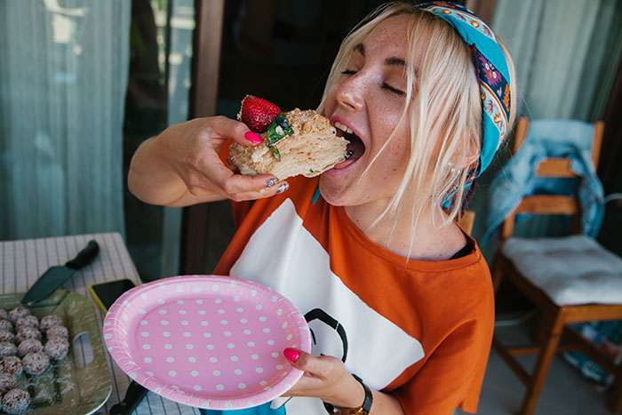 Woman with a headband eating a large slice of peanut butter cake, holding a pink polka dot plate at home.