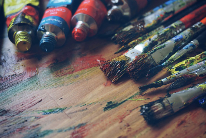 Close up of paintbrushes and paint tubes covered in colorful dried paint on a wooden artist’s workspace surface. - 17