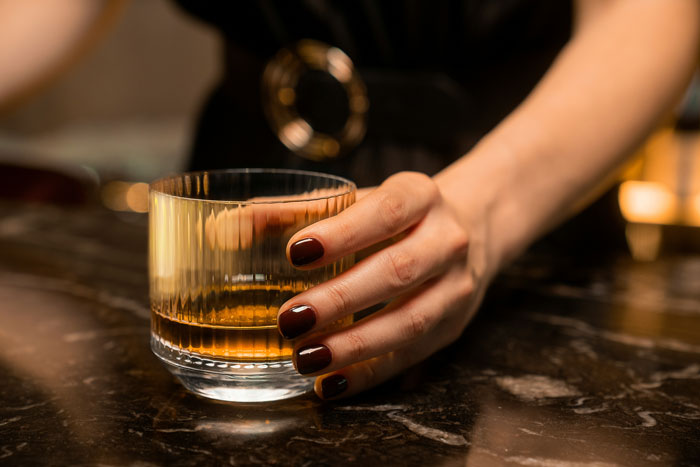 Hand with dark polished nails holding a glass of whiskey on a bar counter, illustrating bar staff protecting women.