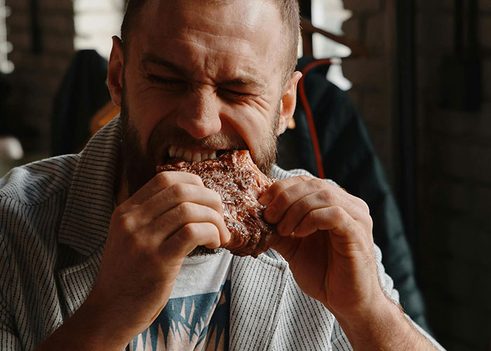 Man biting into a large piece of meat, showing intense focus and enjoyment, reflecting a person tired of fun facts.