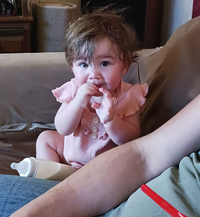 Baby sitting on couch wearing pink outfit with bottle nearby, related to missing mom and baby found case. Baby sitting on couch wearing pink outfit with bottle nearby, related to missing mom and baby found case.