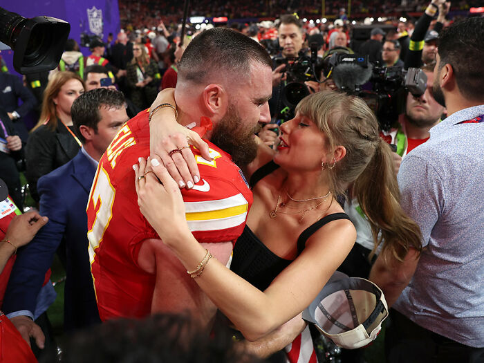 Taylor Swift embracing Travis Kelce on a crowded field with photographers capturing the moment after a game.