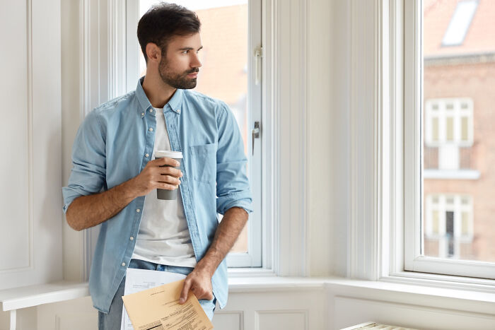 Man holding coffee cup and papers, standing by window, reflecting on smooth pickup lines he might use or has heard.