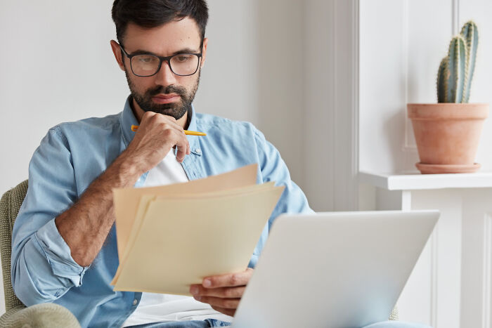 Man with glasses reviewing documents and using laptop at home, focused on unhinged hacks to land job strategies.