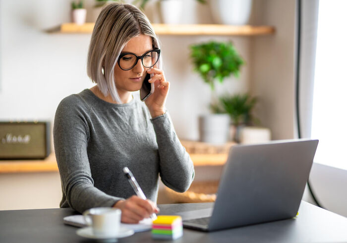 Woman using unhinged hacks to land job, talking on phone and taking notes at a desk with laptop and coffee cup.
