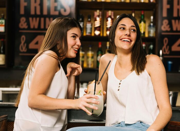 Two women smiling and chatting at a bar, enjoying drinks and sharing smooth pickup lines in a casual setting.