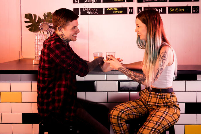 Young tattooed couple smiling and holding hands at a bar, illustrating smooth pickup lines in a casual setting.