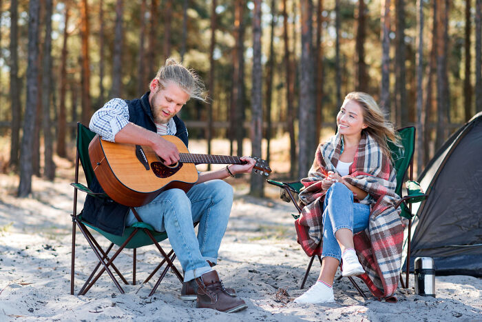 Young man playing acoustic guitar by campfire while woman listens, evoking chilling true stories of scariest sounds heard at night
