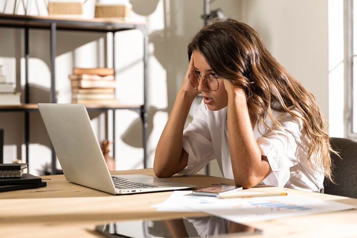 Woman wearing glasses sitting at desk, stressed while looking at laptop, depicting challenges with shopping dependency.