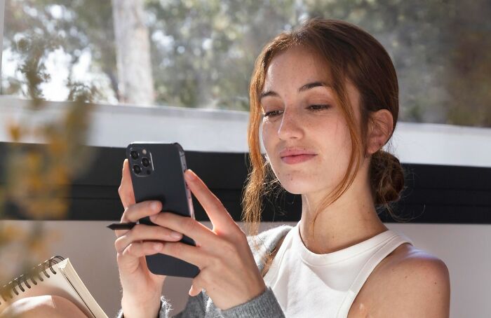 Young woman using smartphone and notebook, focused on unhinged hacks to land job tips indoors by window.