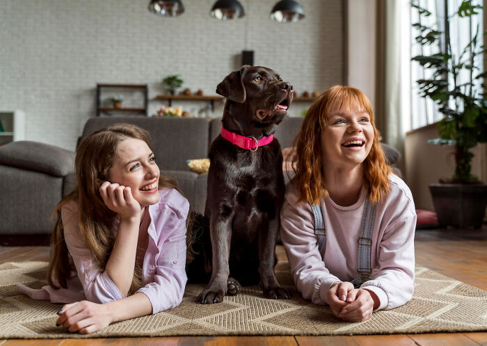 Two women lying on the floor with a black dog between them, smiling and enjoying a relaxed moment indoors.