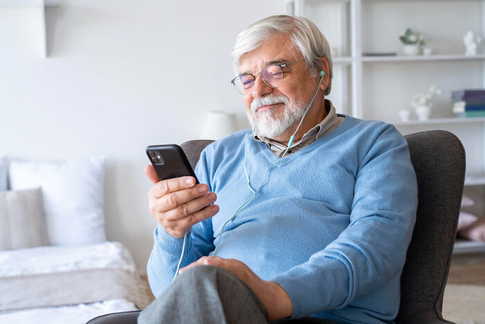 Elderly man using smartphone with earphones, enjoying a low effort high reward moment at home in a relaxed setting.