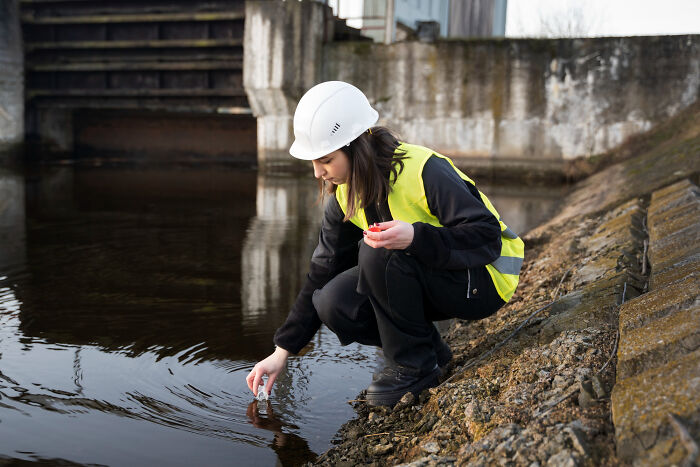 Woman in safety vest and helmet collecting water sample near concrete structure, highlighting aspects of modern life breaking down.