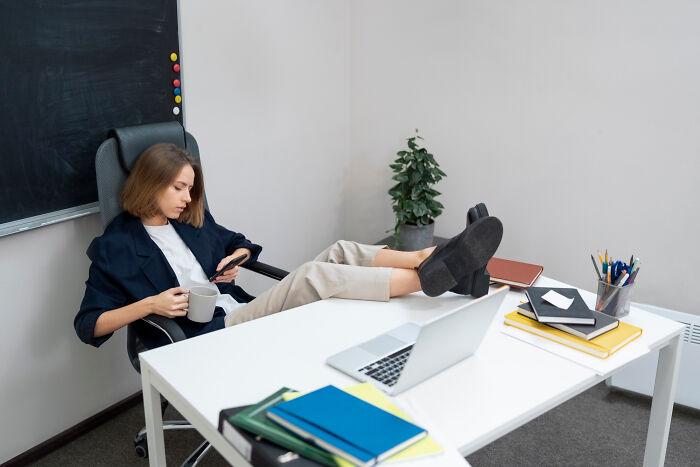 Woman skipping workday, casually sitting with feet on desk, holding coffee and checking phone in office setting.