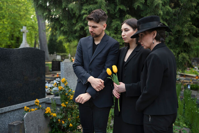 Three employees skipping work day attend a cemetery, dressed in black, holding yellow flowers for an unusual reason.