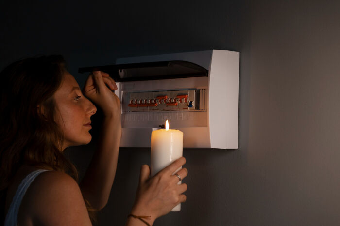 Woman holding a candle while inspecting a circuit breaker, highlighting frighteningly close breakdown of modern life systems.