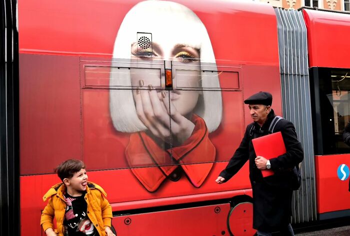 Young boy laughing and man walking near red bus with street art, capturing unseen unposed moments on the streets.