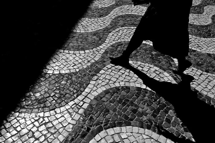 Shadowed legs crossing a patterned cobblestone street, capturing unseen, unposed moments of life on the streets.