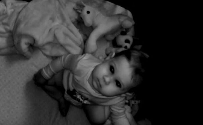 Baby looking up at a home security camera in a crib with stuffed animals, captured in black and white night vision.
