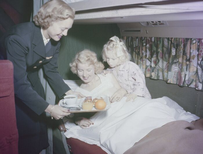 Flight attendant serving breakfast tray to glamorous woman and child in vintage airplane, showcasing flying glamour era.