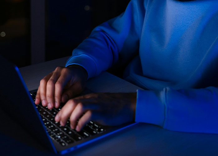 Person wearing a blue sweatshirt typing on a laptop keyboard in a dimly lit room, illustrating drive over the speed limit concept.
