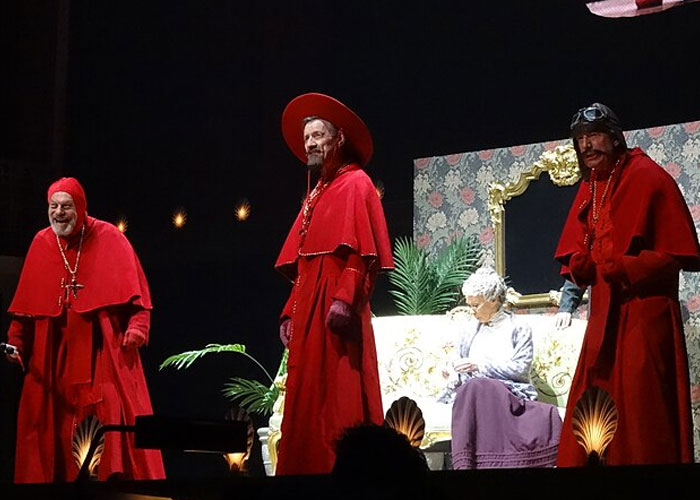 Three men dressed in red cloaks performing on stage in a theatrical play about jobs without a soul.