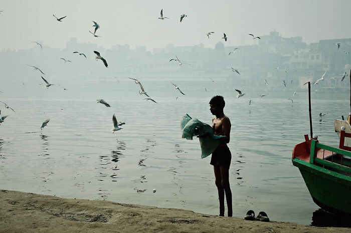 Boy standing by water with birds flying around, capturing candid everyday moments from Asia by photographer Gil Kreslavsky.