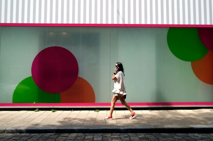Woman walking on sidewalk wearing white dress and red shoes, street moments captured candidly in urban setting with colorful backdrop.