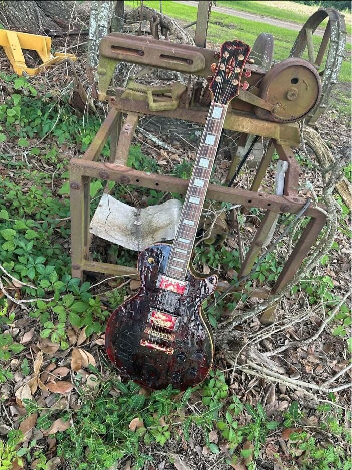 Rusty damaged electric guitar leaning against broken metal frame in overgrown outdoor area, a craft shaming DIY fail.
