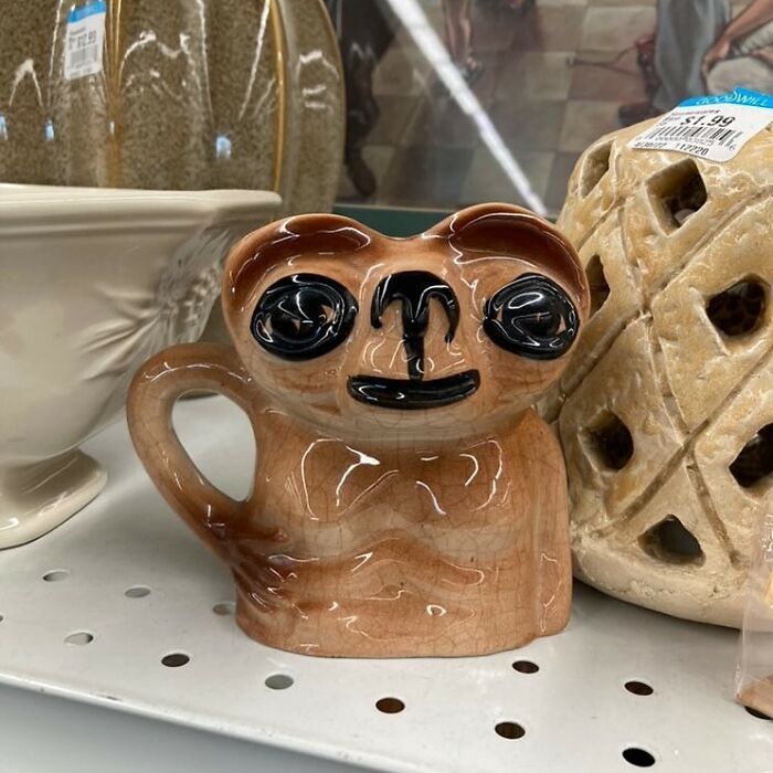 Ceramic mug shaped like a quirky animal face with large eyes, found among bizarre thrift store items on a shelf.