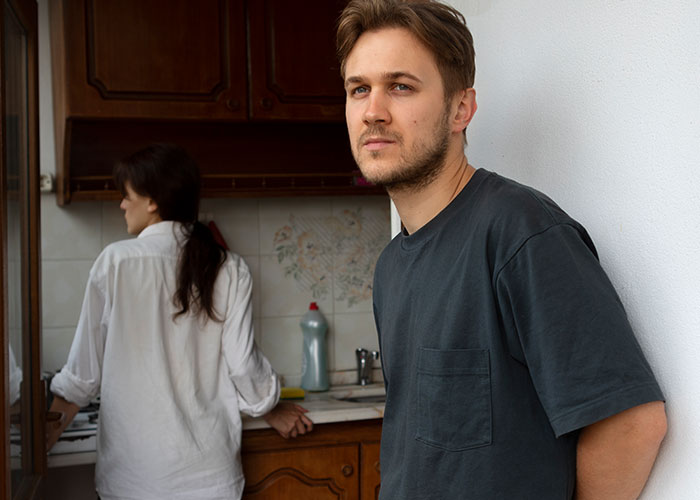 Young man looks worried in kitchen while woman stands near stove, relating to getting sick after eating food dad touched.