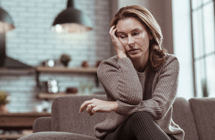 Sad woman sitting on couch in a cozy living room, upset about teen wanting to keep late father last name conflict.